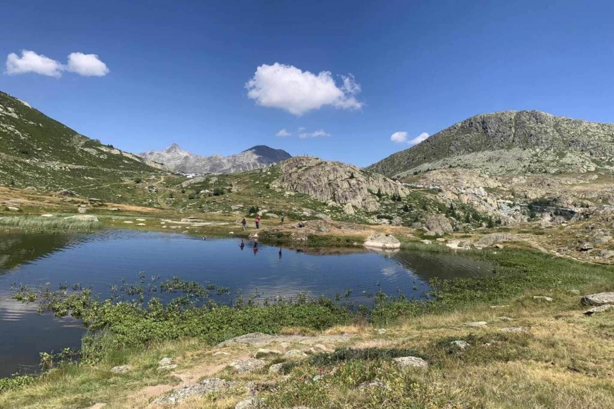 A mountainous landscape with a clear lake, rocky terrain, and a few people in the distance.