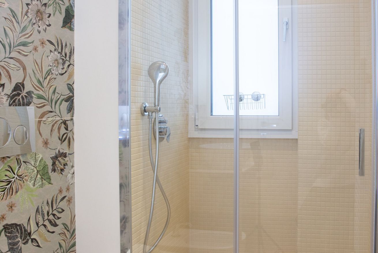 Une douche avec une porte en verre, une fenêtre au-dessus et un mur recouvert de carreaux décorés d’un motif floral.