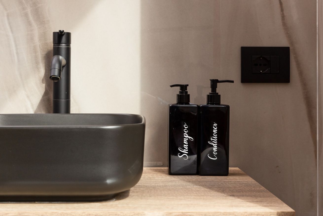 A bathroom with a modern sink, two black bottles of shampoo and conditioner, and a power outlet on the wall.