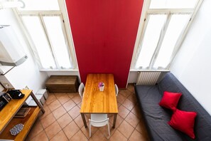 A compact room with a wooden table and chairs, a red accent wall, a grey sofa with red cushions, and a window with curtains. - Casa Modigliani - Porta Romana Apartment (Milano)