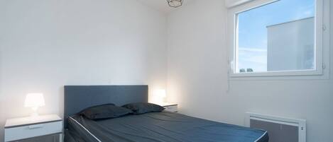 A modern bedroom with a single bed, two bedside tables with lamps, a window with blinds, and a ceiling light fixture.