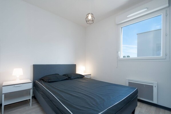 A modern bedroom with a single bed, two bedside tables with lamps, a window with blinds, and a ceiling light fixture.