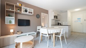 A modern living room with a TV, white furniture, a dining area, and a kitchen in the background.