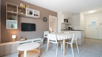 Una sala de estar moderna con televisor, mobiliario blanco, una zona de comedor y una cocina al fondo.