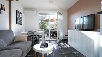 A modern living room with a grey sofa, a white coffee table, and a flat-screen TV on a white cabinet. There is a dining area with a table and chairs, and a sliding glass door leading to an outdoor patio.