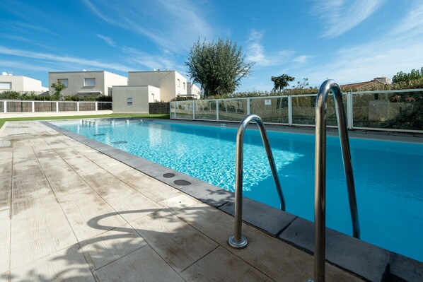 Ein Schwimmbecken mit Holzterrasse und Metallgeländern, umgeben von einem weißen Zaun und Gebäuden im Hintergrund.