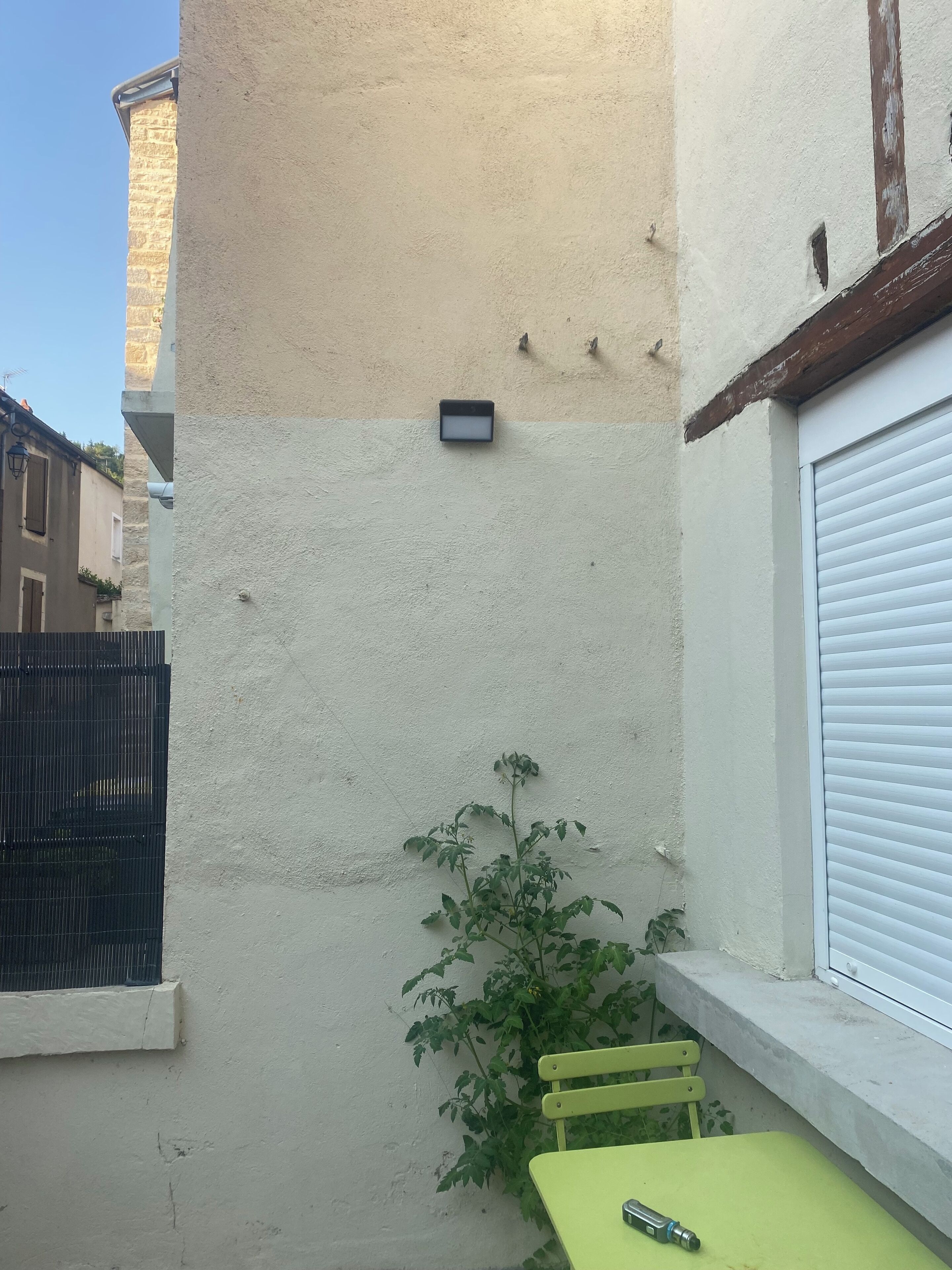 A small outdoor seating area with a table and chairs, a potted plant, and a wall-mounted device.