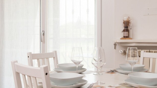 Una mesa de comedor dispuesta con platos blancos y copas de vino, una silla blanca y una ventana con cortinas translúcidas.