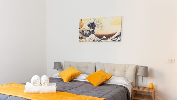 A neatly made bed with a grey and orange bedspread, two pillows, and a pair of rolled towels on the side. - Kandinsky - Exclusive Apartment (Milano)