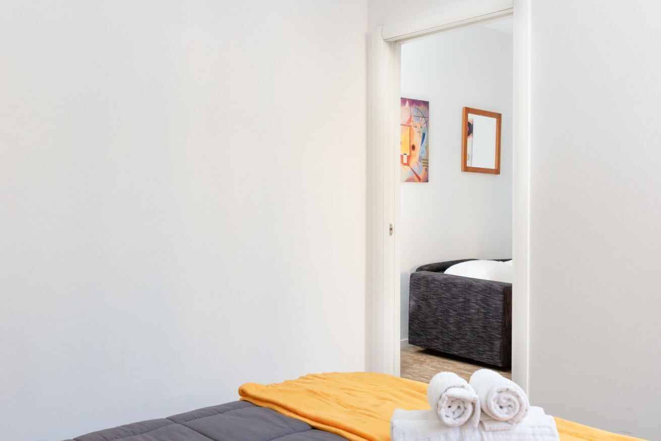 Een hotelkamer met een bed, een houten nachtkastje met handdoeken, een grijze bank en een schilderij aan de muur.