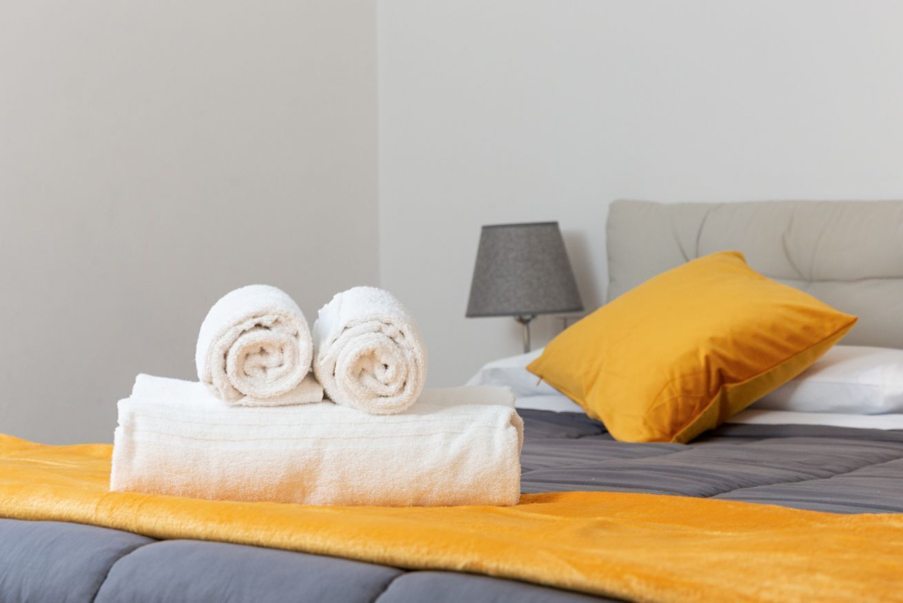 A neatly made bed with a gray and yellow blanket, two rolled white towels, a gray lamp, and a mustard yellow pillow.