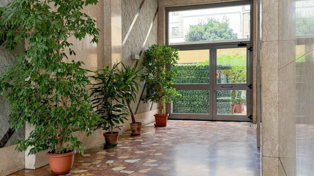 A hallway with a tiled floor, potted plants, and a large glass door leading outside. - Klimt Central station - Two Room Apartment (Milano)