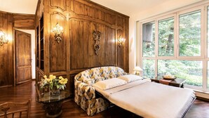 Un dormitorio con un gran armario de madera, una cama con ropa de cama floral, una mesita de cristal con flores y una ventana con vistas a zonas verdes.