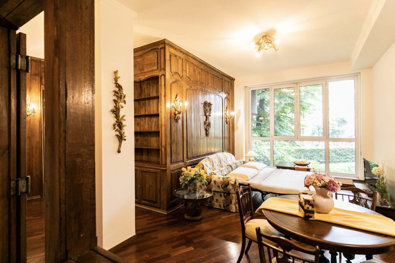 Ein Zimmer mit einem Holzschrank, einem Bett mit blau-weißer Musterdecke, einem Esstisch mit Stühlen und einem Fenster mit Blick ins Grüne.