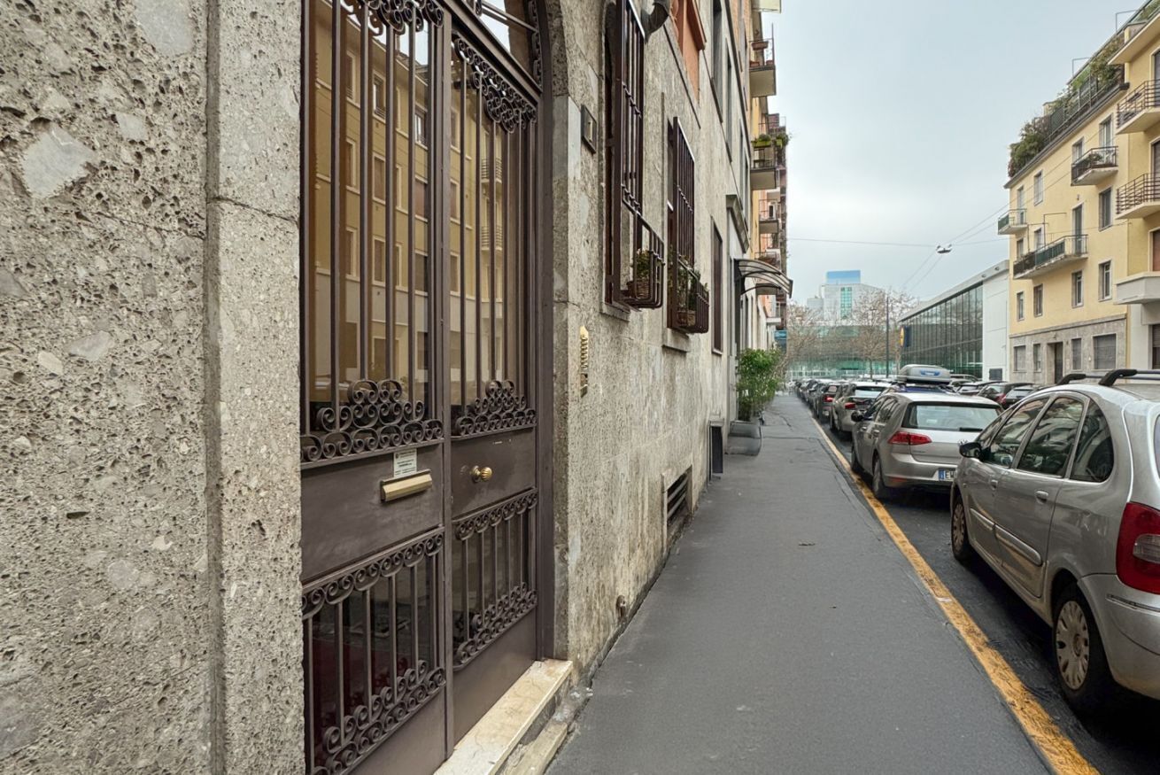 A narrow street with parked cars on one side and a building with barred windows and a door on the other.
