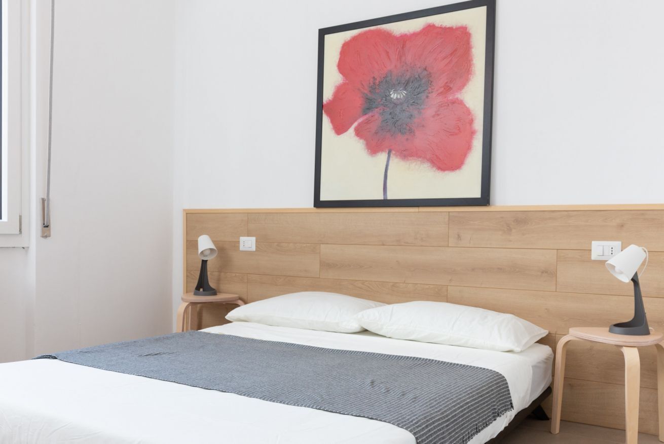 A neatly made bed with white and gray bedding, a wooden headboard, and a framed artwork of a red flower on the wall.