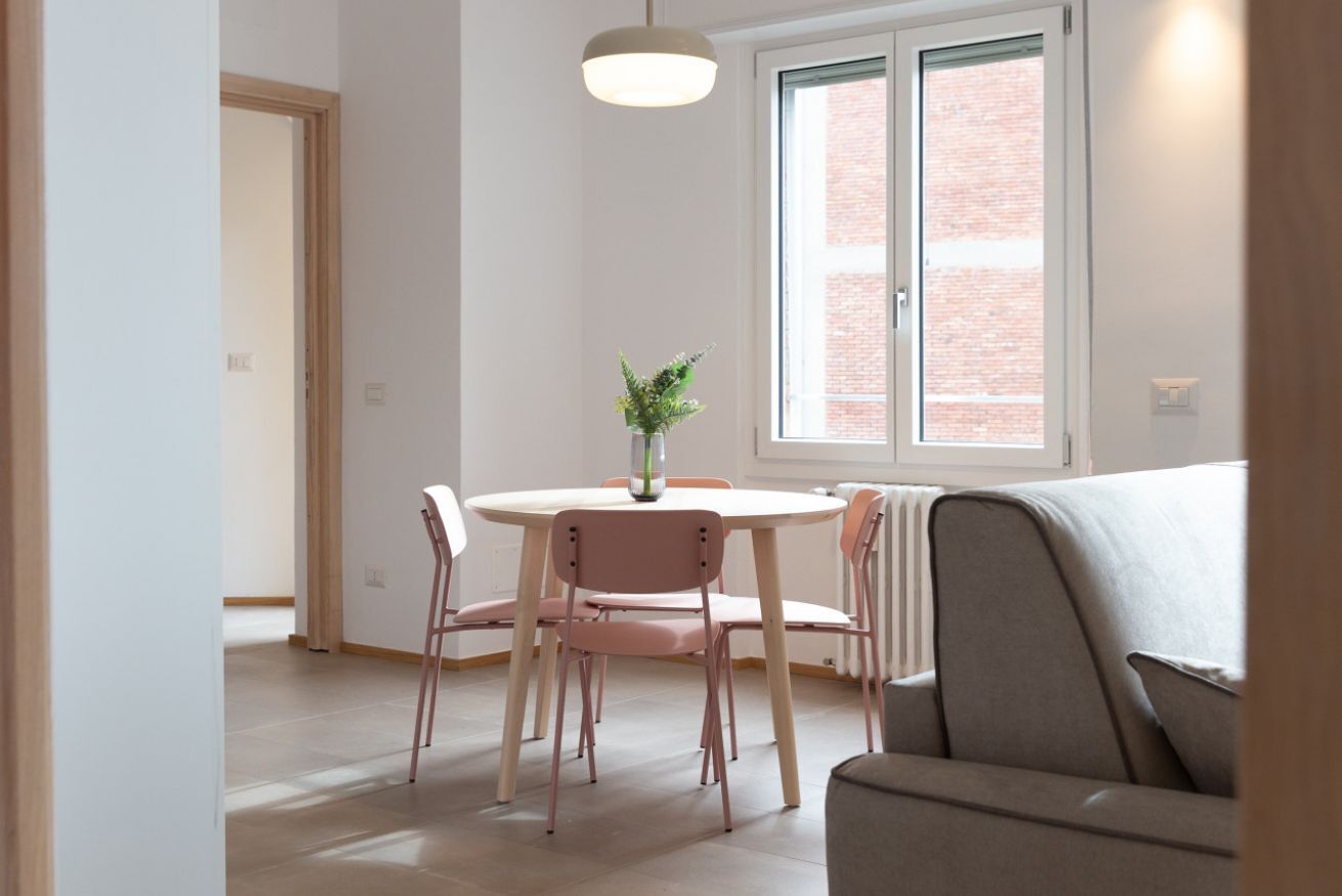 A dining area with a round table, four chairs, a vase with flowers, and a window with a view of a brick building.