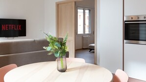 A modern living room with a sofa, a round table with a vase of flowers, a flat-screen TV, and a kitchen area with an oven.