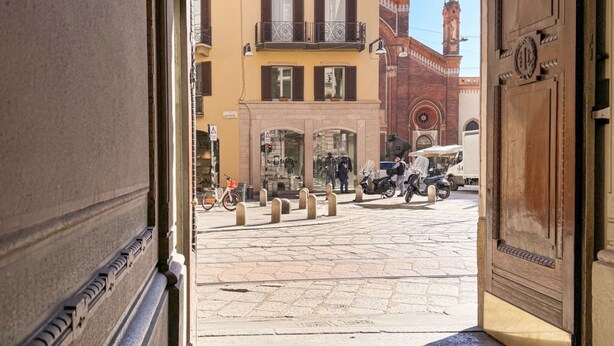 An open doorway leading to a cobblestone street with a building featuring a clock tower and a shop with mannequins. - Piazza del Carmine - Gold Exclusive Apartment - APARTMENT IN THE UNDERGROUND (Milano)
