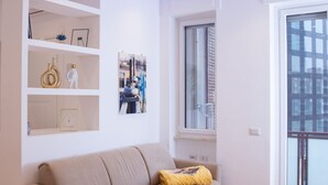 A modern living room with a beige sofa, a small window with a view of buildings, and white walls with shelves holding decorative items.