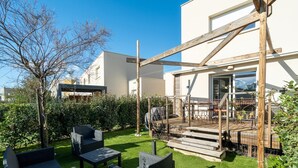Un patio exterior moderno con mobiliario de mimbre, una terraza de madera y vistas a un edificio con grandes ventanales.