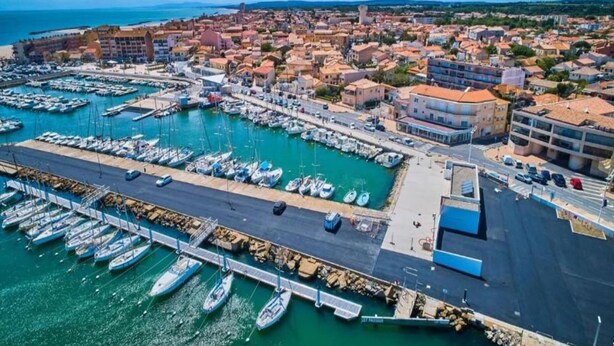 Vista aérea de un pueblo costero con un puerto deportivo, edificios y cielo despejado.