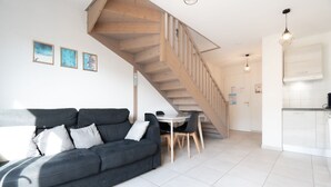 A modern living room with a grey sofa, a wooden staircase, a dining area, and a kitchen with white cabinets. - Charming Villa in Valras-Plage (Valras-Plage)