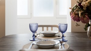 Une table à manger dressée avec deux verres bleus, des assiettes et des couverts, et un vase de fleurs placé sur le côté.