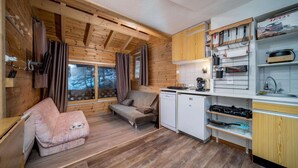 A cozy cabin-style room with a wooden interior, a sofa, a small kitchenette, and a window with curtains. - Apartment in French Alps near Ski Resort (Allos)