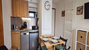 Una cocina compacta con gabinetes de madera, un microondas, una cafetera y un lavavajillas. Hay una mesa de comedor con sillas y un reloj en la pared.