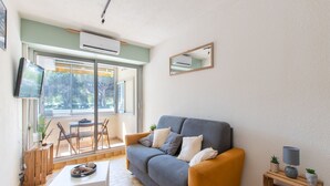 A living room with a grey sofa, a white coffee table, and a small wooden side table with a lamp. There is a TV mounted on the wall, a window with a view of trees, and a sliding glass door leading to an outdoor area.