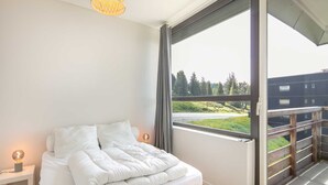 A bedroom with a large window, a bed with white bedding, a bedside table with a lamp, and a view of a building and greenery outside. - Alpine Escape at Chamrousse (Chamrousse)
