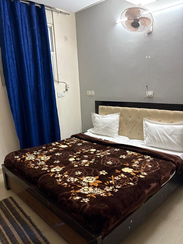 A bedroom with a bed, a wall-mounted fan, a power outlet, and a window with blue curtains. - ICY WHITE STAY HERE (Noida)