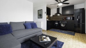 Una sala de estar moderna con un sofá gris, una mesa de centro con controles remotos, y una cocina con lavadora, microondas y armarios negros.
