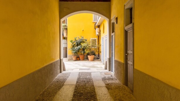 A narrow alleyway with yellow walls and a stone floor, leading to a courtyard with potted plants. - Located on the second floor of a typical Milanese condominium, this two-room apartment combines comfort and contemporary style. The flat features a fully equipped kitchen, ideal for those who love to cook and host. You will find a hob, oven, fridge, dish (Milano)