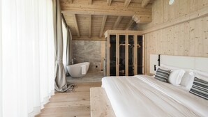 Une chambre avec un plafond en bois, un grand lit avec une literie blanche, un banc et un dressing avec des portes en verre.