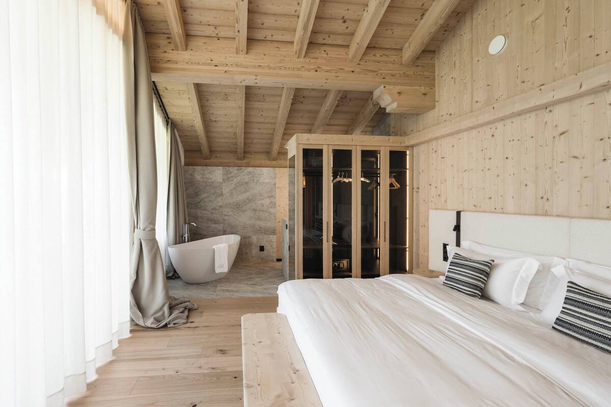 A bedroom with a wooden ceiling, a large bed with white bedding, a bench, and a walk-in closet with glass doors.