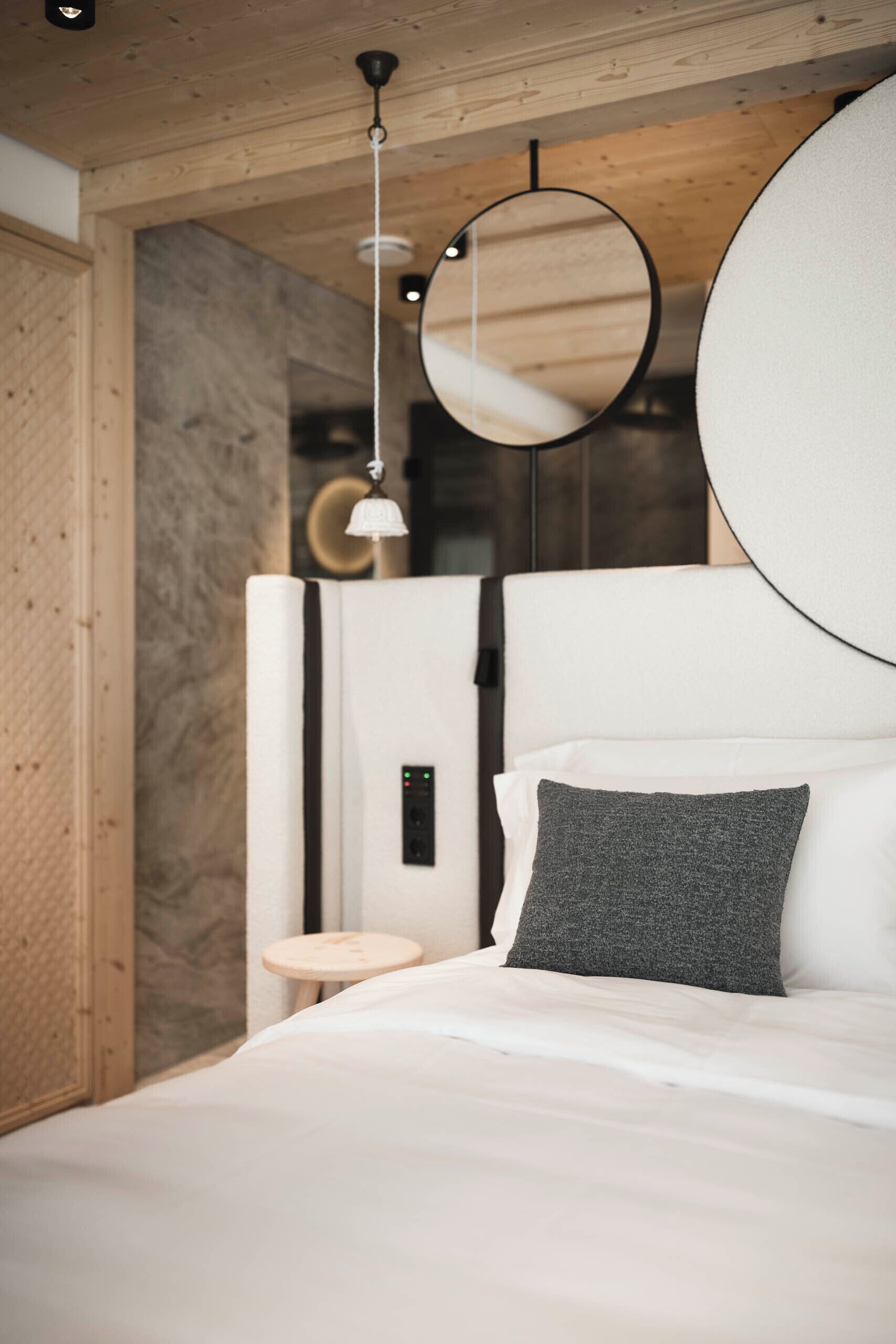 A modern bedroom with a wooden ceiling, a round mirror, a bedside table, and a bed with white and gray bedding.