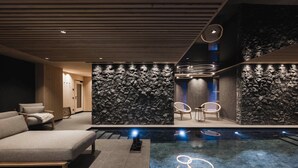 Un espace piscine intérieur moderne, avec un mur en pierre, des chaises longues et un coin salon comprenant une table et des chaises.