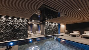 Un espace piscine intérieur moderne, doté d’un mur en pierre, de chaises longues et d’un coin salon avec des canapés.