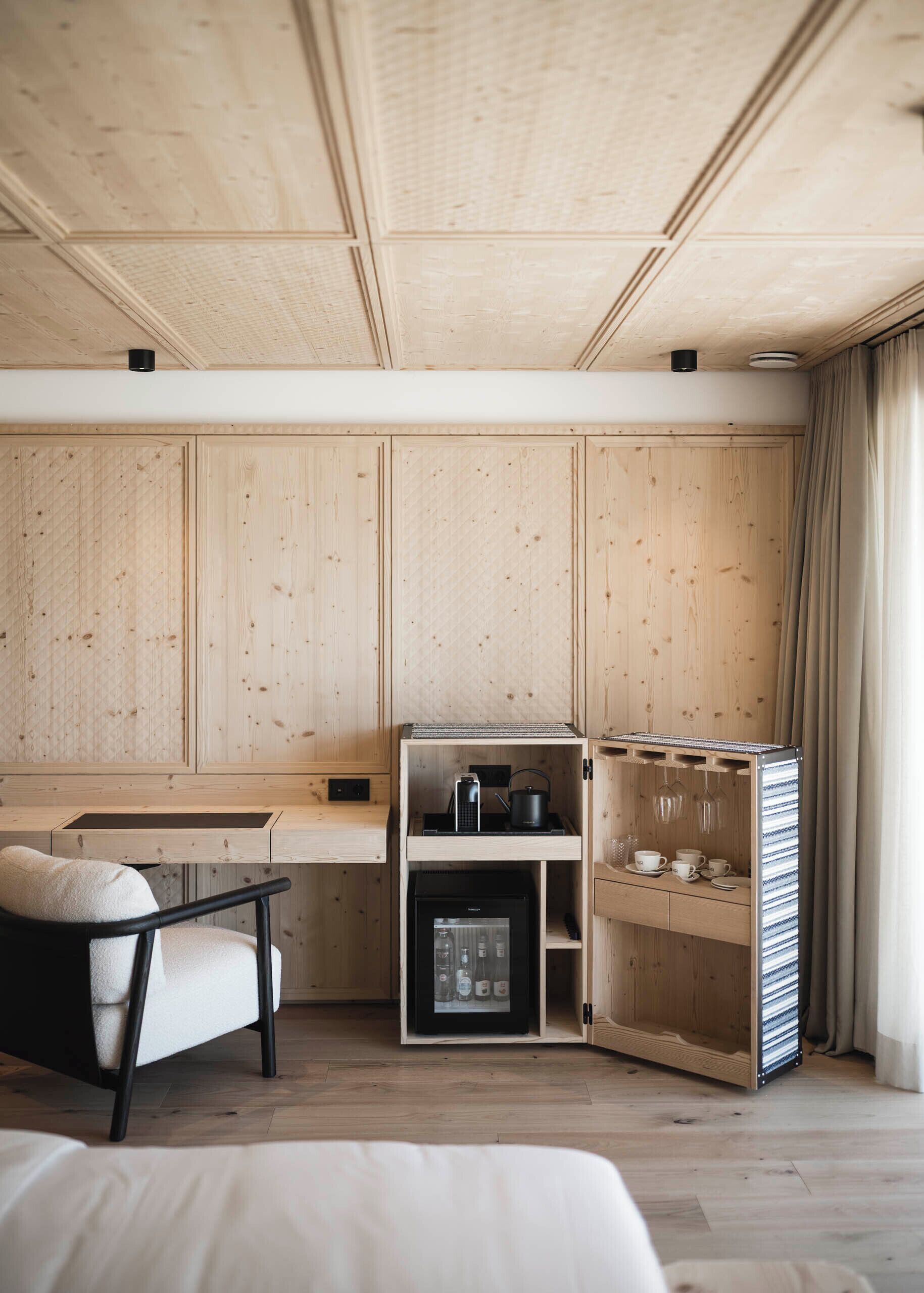 A compact, modern room with wooden panel walls, a small kitchenette, a seating area with a sofa and a table, and a cabinet with storage shelves.