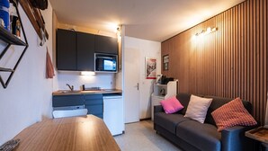Ein kompakter Wohnraum mit Küchenzeile, einem Esstisch, einer Couch mit Kissen und einer Wand als Holzoptik.