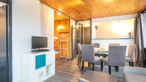 A modern room with a wooden ceiling, a flat-screen TV on a white cabinet, a dining area with grey chairs, and a small table. - Alpine Comfort at Chamrousse (Chamrousse)