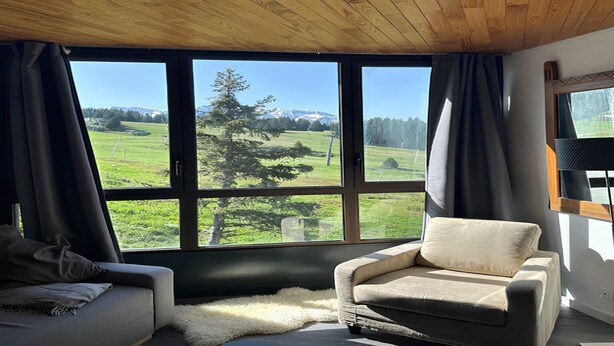 A room with a large window offering a view of a green landscape and mountains. There is a sofa and a chair inside the room. - Alpine Comfort at Chamrousse (Chamrousse)
