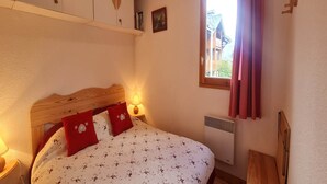 A bedroom with a wooden headboard, a bed with floral bedding, a nightstand with a lamp, a window with a view of buildings, and a radiator.