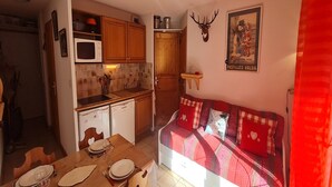 A compact kitchen with wooden cabinets, a microwave, and a sink. There is a dining table set with plates and glasses. A bed with a red and white checkered blanket is visible.