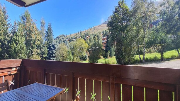 A wooden deck with a table and chairs, overlooking a mountainous landscape with trees and houses.