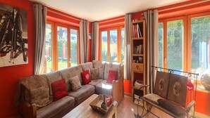 Una sala de estar acogedora con un sofá seccional, una mesa de centro de madera y una estantería. Amplias ventanas con cortinas dejan entrar luz natural y ofrecen vistas al exterior.