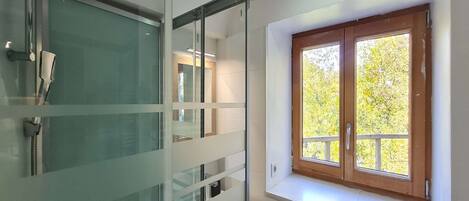 Ein Badezimmer mit Duschkabine aus Glas, einem Fenster mit Blick auf Bäume und einem weißen Waschbecken.