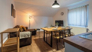 Ein gemütliches, gut beleuchtetes Loft-Apartment mit Essbereich, einer Couch, einem kleinen Tisch und einem großen Fenster mit Jalousien.